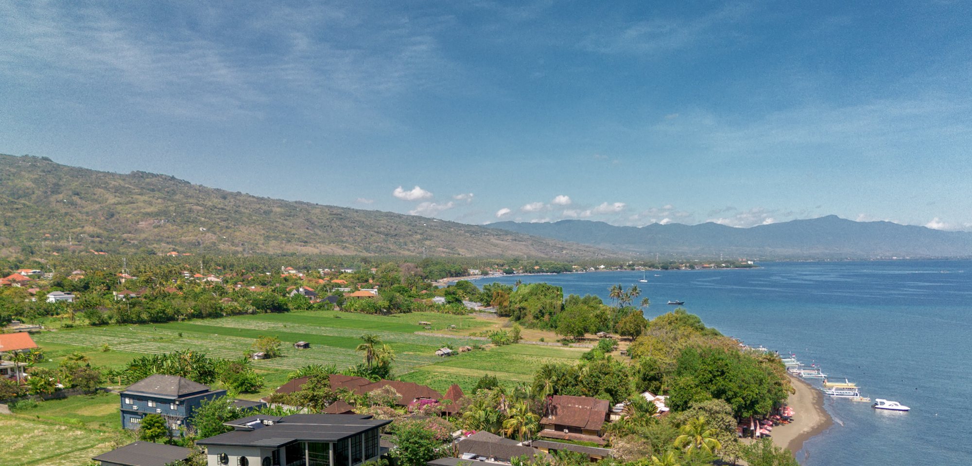 The north Bali coast from the air near Lovina: rice fields and villages under the mountain ridge, the shoreline curving away to moored boats and the open sea